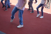 Zu sehen sind die Beine vieler Kinder, die gemeinsam auf einem Sportplatz einen Tanz einüben