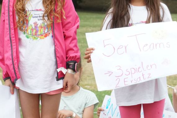 Zwei Mädchen halten ein Plakat hoch, auf welchem steht: 5er Teams, 3 spielen, 2 Ersatz.
