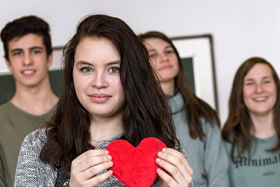 Eine Schülerin hält ein Papierherz vor ihre Brust. Hinter ihr stehen drei weitere Schüler*innen.