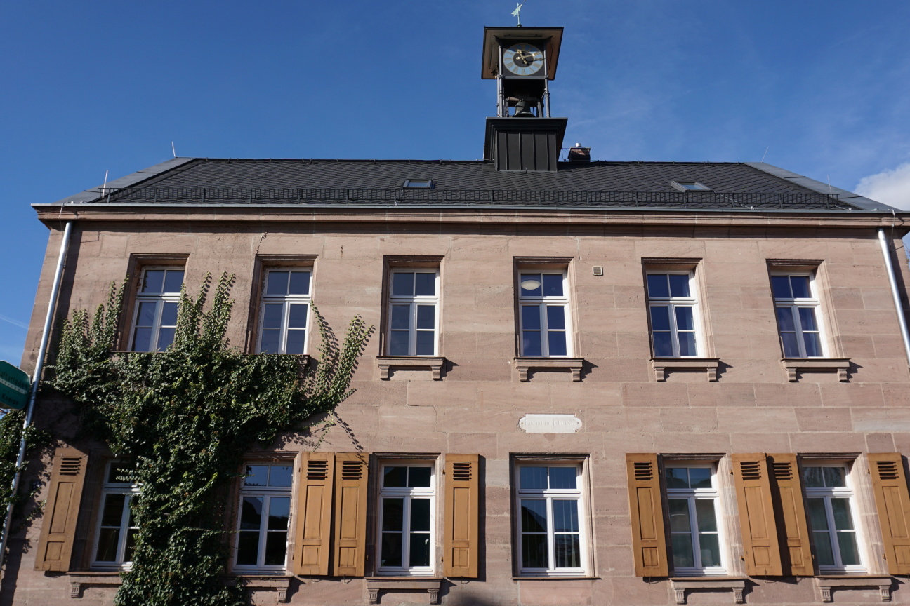Über einem zweistöckiges Sandsteinhaus mit Turmuhr auf dem Dach leuchtet blauer Himmel