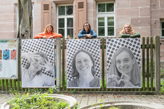 Vor einem alten Sandsteinhaus, sind die Mitarbeiterinnen des Kulturlades Röthenbach zu sehen, wie sie hinter schwarz weißen Plakaten stehen. Die Plakate zeigen ihre Gesichter in Großformat.