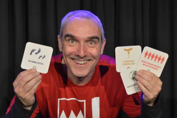 Ein Mann mit rotem T-Shirt hält vier farbige Bierdeckel in den Händen und grinst in die Kamera.