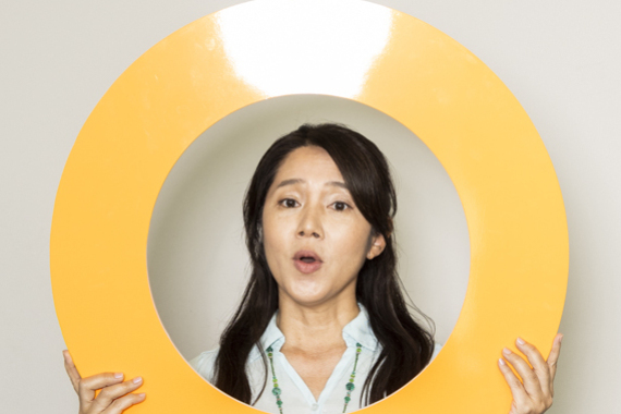 Eine junge Frau mit langen schwarzen Haaren und einem weißen Oberteil schaut durch einen gelben Ring in die Kamera.