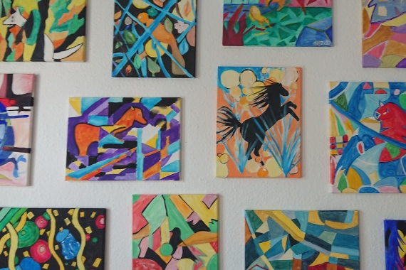 An einer Wand hängen 14 abstrakte Bilder in bunten Farben