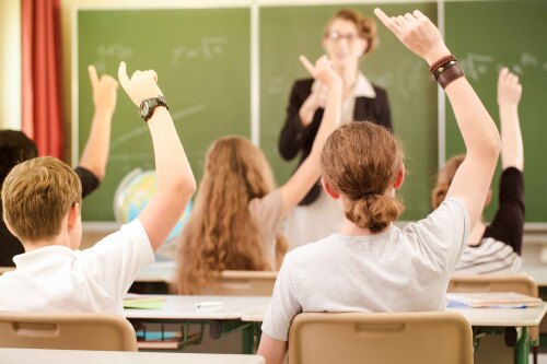© Kzenon / AdobeStock Schüler melden sich im Unterricht. Eine Lehrerin steht vor einer Tafel. © Kzenon / AdobeStock