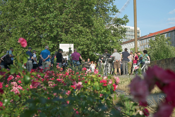 Blumen im Vordergrund; im Hintergrund haben sich Menschen bei einer Mobilen Bürgerversammlung versammelt.