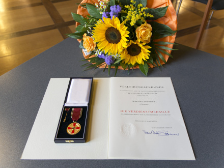 Das Bild zeigt eine Ehren·urkunde und eine Medaille auf einem Tisch. Auf dem Tisch liegt auch noch ein bunter Blumen·strauß.