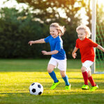 Kids play football on outdoor field. Children score a goal at soccer game. Girl and boy kicking ball. Running child in team jersey and cleats. School football club. Sports training for young player.