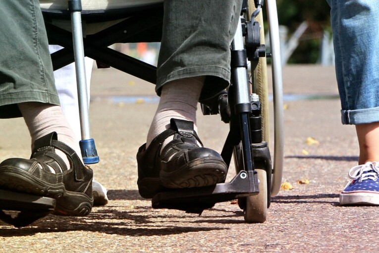 Nahaufnahme von den Füßen/Rädern einer Person im Rollstuhl und einer Person daneben mit bunten Turnschuhen.