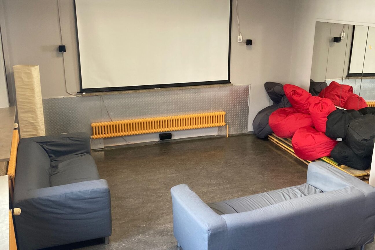 Zwei graue Sofas stehen vor einer weißen Leinwand. An der Decke hängt ein Videobeamer. Im Hintergrund sind rote und schwarze Sitzsäcke aufgestapelt., Bild © Jugendtreff MAX