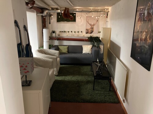 Im Kellerraum steht ein weißes Sofa auf einem dunkelgrünen Teppich. An der Decke sind Heizungsrohre, an der Wand hängen zwei kleine blaue Ukulelen. © Jugendtreff MAX