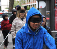 Ein junge mit Augenbinde geht durch die Straße. Ihm folgen weitere Kinder.