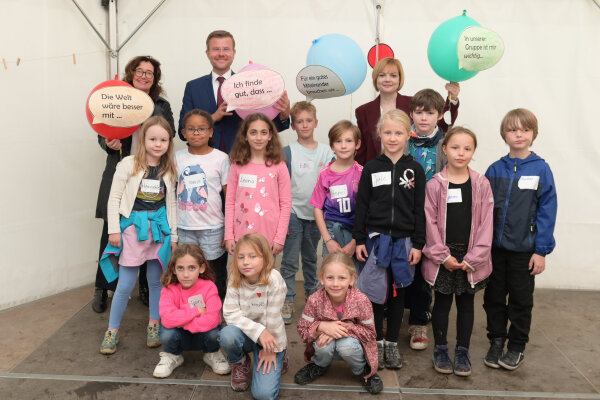 In der letzten Reihe stehen Anekatrin Fries, Markus König und Jana Stadlbauer und halten Luftballons in die Höhe auf denen Sprechblasen mit Satzanfänge wie "Die Welt wäre besser mit..." geklebt sind. Davor stehen und sitzen mehrere Jungen und Mädchen in zwei Reihen.