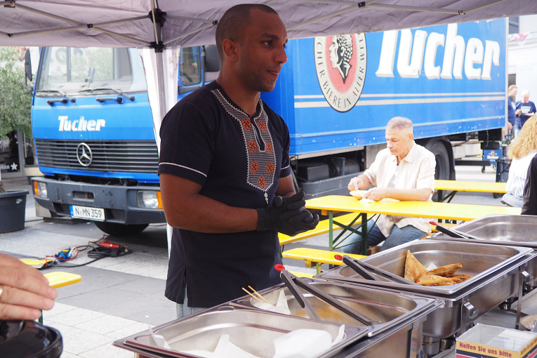 Ein Man der Essen austeilt am Friedenstafel Publikum mit dem Tucher Wagen im Hintergrund