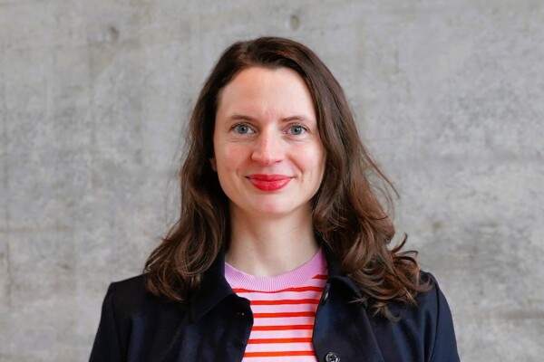 Frau mit rosa rotem Oberteil steht vor grauem Hintergrund und lächelt in die Kamera