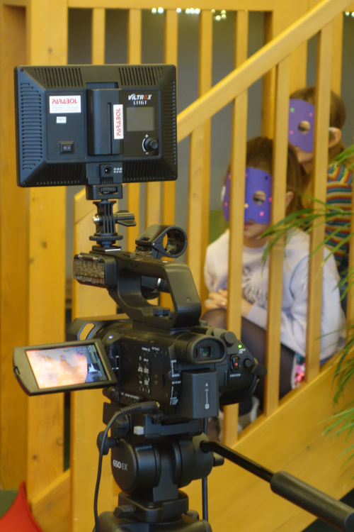 Im Vordergrund steht eine Videokamera. Zwei Kinder sitzen dahinter auf der Treppe und beobachten den Videodreh.