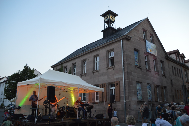 Vor einem Sandsteinhaus mit golden glänzender Turmuhr steht eine Bühne, auf der eine Band spielt.