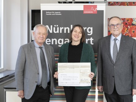3 Personen stehen vor einem großen Poster von Nürnberg engagiert. Die Frau in der Mitte ist die Sozial·referentin Elisabeth Ries von der Stadt Nürnberg. Frau Ries hält einen großen Spenden·scheck in der Hand.