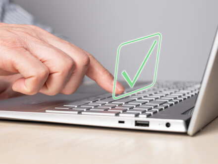 Das Bild zeigt einen aufgeklappten Laptop. Eine Hand streckt einen Finger aus und drückt eine Taste auf der Tastatur. Über der Tastatur sieht man einen grünen Haken. Der Haken ist das Symbol für: Bestätigen.