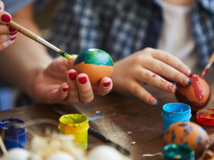 Das Bild zeigt Hände von einer erwachsenen Person und einem Kind. Beide Personen malen Oster·eier mit Farbe und einem Pinsel an.