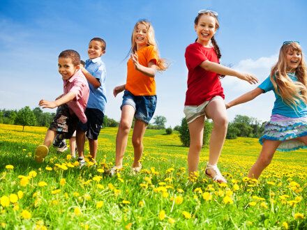 Das Bild zeigt 5 Kinder. Die Kinder rennen über eine Wiese und lachen. Das Wetter ist schön.