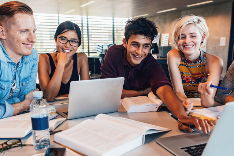 Das Bild zeigt 5 junge Personen beim gemeinsamen Lernen. Die Personen sitzen an einem Tisch. Auf dem Tisch sind ein Laptop und viele Bücher. Die Personen lächeln.