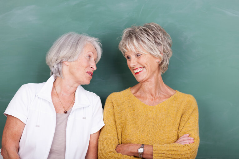 Das Bild zeigt 2 ältere Frauen. Die Frauen stehen nebeneinander vor einer Schul·tafel. Die Frau links sagt etwas zur Frau rechts. Die Frau rechts lächelt.