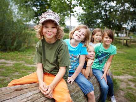 Das Bild zeigt 5 Kinder. Die Kinder sitzen auf einem Baum·stamm. Die Kinder sind unterschiedlich alt.