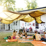 Viele Menschen sitzen auf einer Wiese unter einem Stoffdach vor den Pavillons der Kunstakademie.