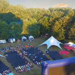 Luftaufnahme von vielen Stühlen und Zelten, die vor einer Open-Air-Kinoleinwand auf einer Wiese stehen