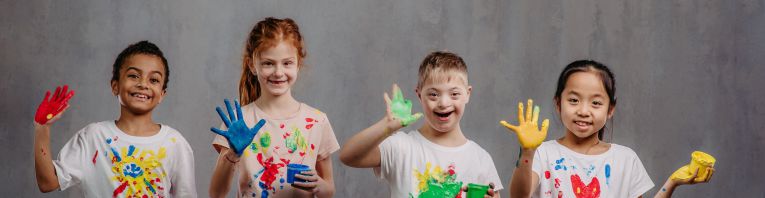 Vier Kinder mit bemalten Händen und T-Shirts stehen lachend mit Fingerfarben in der Hand nebeneinander