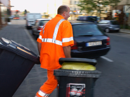 Das Bild zeigt einen Mitarbeiter von der Müll·abfuhr. Der Mitarbeiter zieht zwei Müll·tonnen hinter sich her.