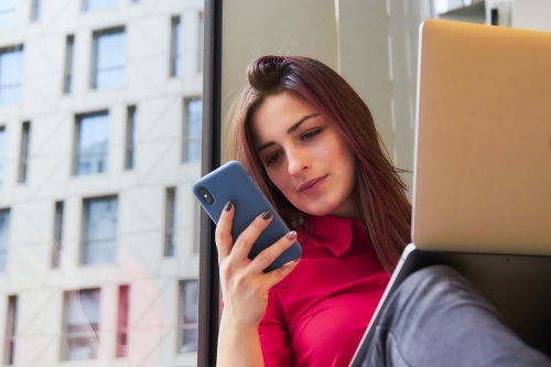 Eine Frau sitzt an einem Fenster mit einem aufgeklappten Laptop auf den Knien und einem Handy in der Hand.