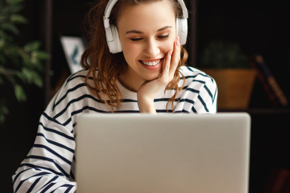 Eine Person sitzt an einem Schreibtisch an einem Laptop. Sie trägt weiße Kopfhörer, blickt auf den Bildschirm und lächelt.