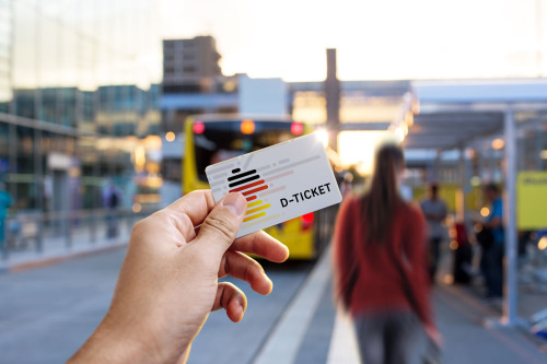 Eine Hand hält ein symbolhaft gestaltetes Deutschlandticket in die Luft. Im Hintergrund ist der Bahnsteig eines großen Bahnhofs zu sehen.