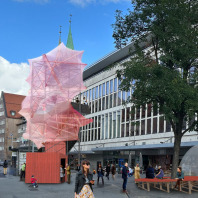 Ansicht der Fassade des Galeria-Gebäudes in der Königstraße in Nürnberg. Davor befindet sich die Illustration einer meterhohen Kunstinstallation bestehend aus einem Gerüst, das mit einem rosafarbenen Tuch umspannt ist.