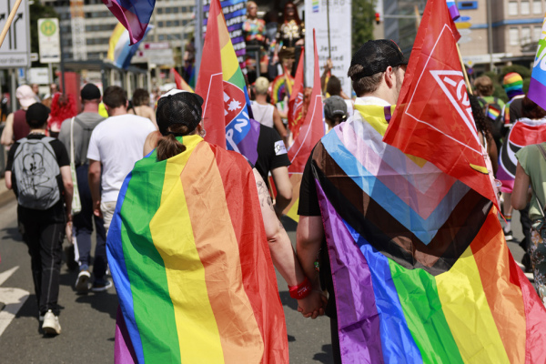 CSD Demo in Nürnberg im Jahr 2024: Zwei Teilnehmer sind von hinten zu sehen. Sie tragen beide einen Umhang in Regenbogenfarben. Menschen halten Regenbogenfahnen über ihren Köpfen in die Luft.