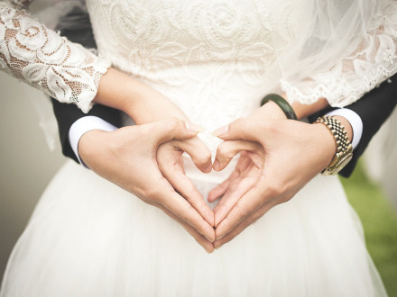 Das Bild zeigt ein Braut·paar. Der Mann und die Frau halten sich an den Händen und machen mit den Fingern ein Herz.
