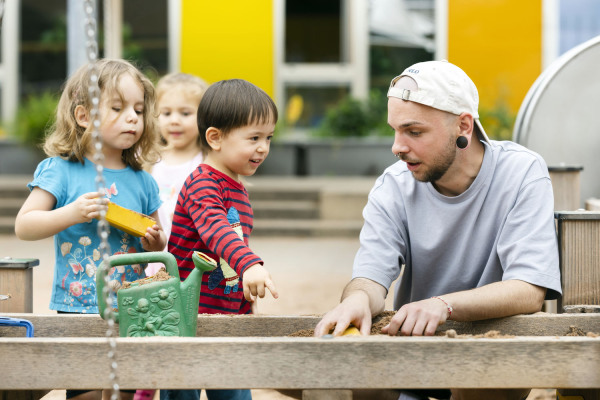 Ein Erzieher aus einer Nürnberger Kita spielt mit Kindern im Sandkasten.