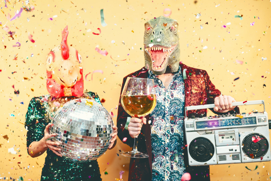 Ein Mann und eine Frau, verkleidet mit einer Dino-Maske und einer Hahn-Maske halten eine Diskokugel und einen Ghettoblaster. Lametta rieselt von der Decke.