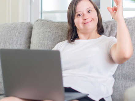 Das Bild zeigt eine junge Frau mit einem Laptop. Die junge Frau sitzt auf einem Sofa. Die junge Frau macht mit der linken Hand das Zeichen für OK.
