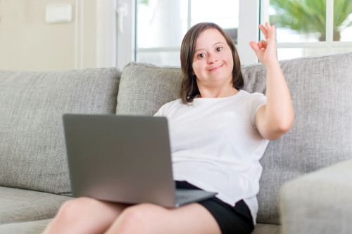 Eine Frau mit Down Syndrom sitzt zuhause auf dem Sofa und hat einen Laptop auf dem Schoß.
