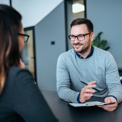 Mann mit Clipboard spricht mit Frau am Besprechungstisch. © AdobeStock