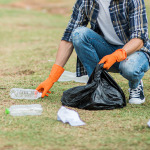 Eine Person mit einem schwarzen Müllsack in der linken Hand befindet sich in der Hocke auf einem Rasen und sammelt leere Plastikflaschen ein. Die Person trägt Chucks, eine Jeans, ein weißes T-Shirt und darüber ein kariertes Hemd.