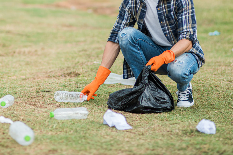 Eine Person bückt sich, um eine leere Plastikflasche von einer Wiese aufzuheben. In der linken Hand hällt sie einen Müllsack. Sie trägt Chucks, Jeans, ein weißes T-Shirt, ein Karohemd und orangefarbene Gummihandschuhe.