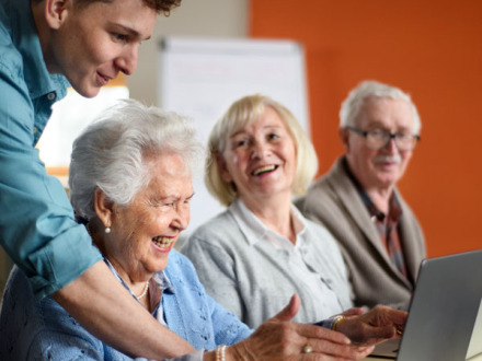 Das Bild zeigt 3 ältere Personen vor einem Laptop. Eine jüngere Person erklärt etwas.