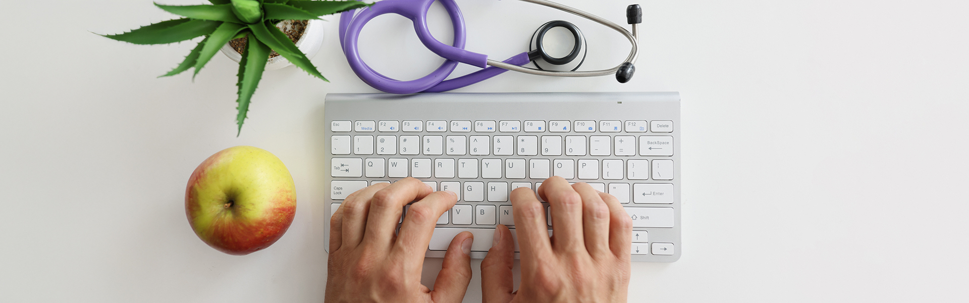 Vogelperspektive, ein Apfel, eine Tastatur mit Händen darauf und ein Stethoskop