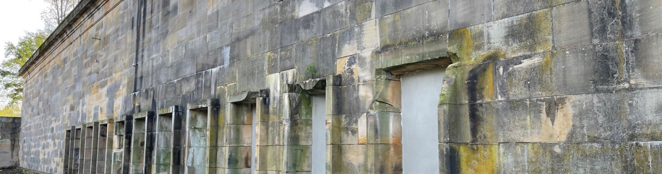 Bild © Andreas Franke Fassade des Bahnhofs Märzfeld. In die Sandsteinmauer sind zehn Türen eingelassen. Die modrige Mauer ist im vorderen, oberen Bereich brüchig und im Vordergrund und Hintergrund von Grün umgeben., Bild © Andreas Franke