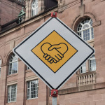 Dieses Foto der Aktion „Verkehrsschilder der Gerechtigkeit" ist während der Nürnberger Wochen gegen Rassismus 2022 entstanden. Es zeigt ein Vorfahrtszeichenschild bestehend aus einem auf die Spitze gestelltem Quadrat. Es ist in der Mitte gelb und außen weiß umrandet. Anders als bei echten Verkehrsschildern befindet sich im gelben Bereich eine Grafik, die zwei Hände zeigt, die sich die Hand geben.