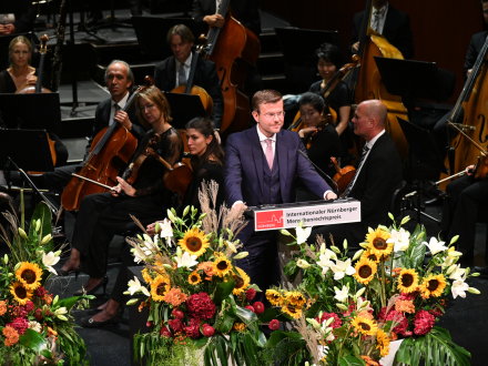 Das Bild zeigt den Nürnberger Ober·bürgermeister Marcus König bei der Verleihung vom Internationalen Nürnberger Menschen·rechts·preis auf der Bühne. Hinter dem Ober·bürgermeister sieht man Musiker aus einem Orchester. Vor dem Ober·bürgermeister stehen sehr viele bunte Blumen.